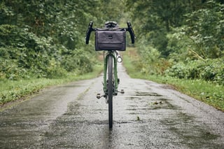Front view of the Woodland Green Faran 2 with a Gramm Tourpacking handlebar bag, on a wet tree-lined lane.