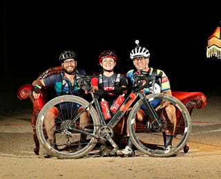 Three smiling riders in helmets sit on a red sofa behind a mud-covered gravel bike at night.