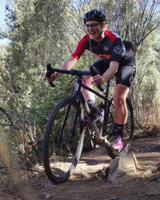 Rider in red and black kit grins while riding a gravel bike down a rocky dirt trail past scrub.