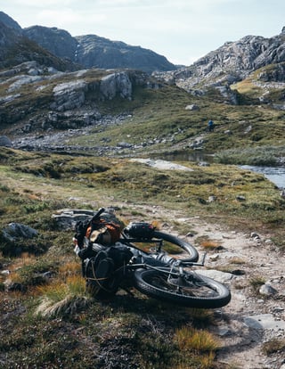 Loaded bikepacking bike lying on a rocky trail by a stream, with a rider climbing the hillside beyond.