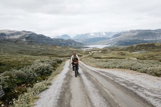 Bikepacker rides a wide gravel road across open Norwegian highland with a lake and mountains ahead.