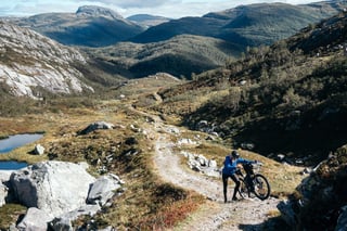 Rider pushes a loaded bike up a stony mountain track in Norway, with lakes and ridges behind.