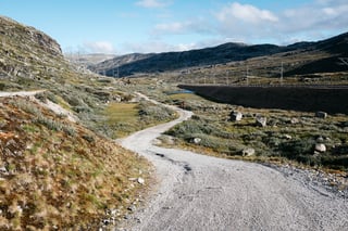Winding gravel track through a bare Norwegian valley, with a distant rider and power lines on the ridge.