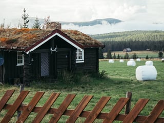 Dark log cabin with a turf roof and wrapped hay bales in a green field, with forest beyond.