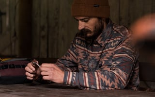 A bearded man in a Cascada beanie and patterned shirt works on a small item at a table beside a bag.