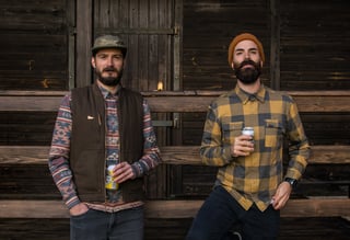 Two men in Cascada flannel shirts and hats stand against a wooden barn, each holding a can.