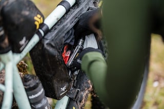 Rider pulls a multi-tool from an Apidura Backcountry frame pack on a muddy mountain bike.