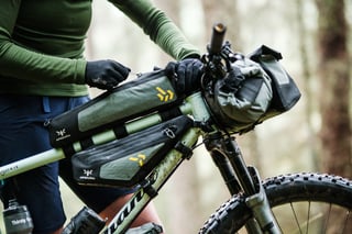 Rider unzips an Apidura Backcountry top tube pack above a frame pack, with handlebar pack fitted.