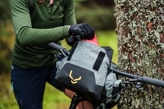 Rider reaches into an open Apidura Backcountry handlebar pack, the bike leant against a lichen-covered tree.