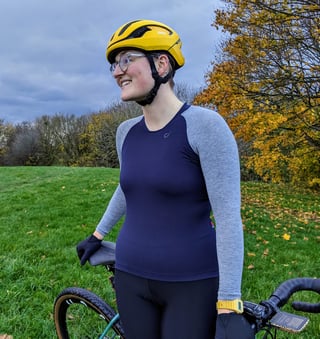 Rider smiling in the Velocio Micromodal long sleeve jersey, standing with a gravel bike on an autumn hillside.