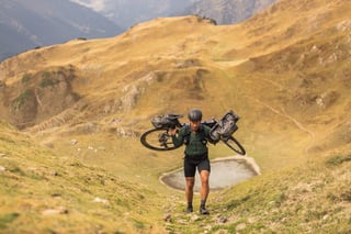 Rider shoulders a loaded gravel bike up a steep grassy slope above a small mountain tarn in the Pyrenees.