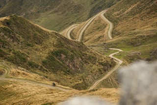 A lone bikepacker rides a hairpin gravel track winding across steep grassy mountainsides in the Pyrenees.
