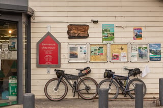 Two loaded gravel bikes lean against a wooden-clad village shop front hung with French signs and noticeboards.