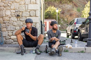 Svein Tuft and Christian Meier sit on a village kerb eating from bowls, helmets on, with beer and water bottles beside them.