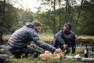 Two friends in down jackets and beanies share bread and wine on the grass beside a river at an evening camp.