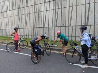 Four cyclists paused with their bikes on a road beside a high concrete wall, one bent over a bike fitted with a pannier.