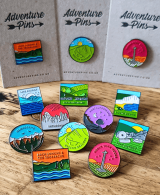 Adventure Pins enamel pins of UK national parks, including Cairngorms, Snowdonia and Brecon Beacons, on a wooden table.