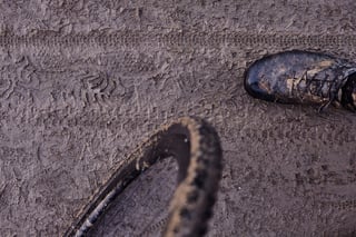 Gravel wheel and a muddy shoe beside deep tyre and boot prints in wet, grey mud on a Ravager test ride.