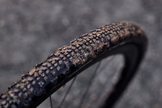 Mud-clogged Maxxis Ravager tread on a gravel wheel, with small square knobs on a blurred grey surface.