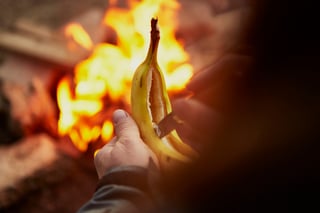 Hands slit a banana lengthways in front of campfire flames, preparing a banana boat with Outdoor Provisions snacks.