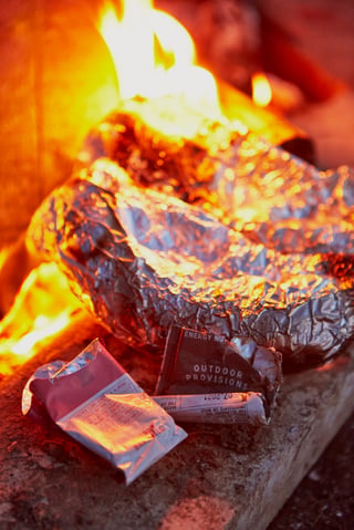 Foil-wrapped banana boat cooking in campfire flames, with empty Outdoor Provisions wrappers on the stone below.