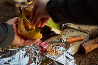 Banana filled with Outdoor Provisions bar crumbs held over foil, with a knife and bar wrapper beside the fire.