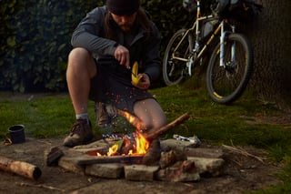 Rider in a beanie cuts into a banana beside a campfire, a loaded bikepacking bike leaning on a tree behind.