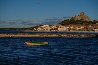 Ruined hilltop tower above a lagoon-side village, with a yellow and red fishing boat moored in the foreground.