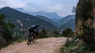 Bikepacker on a loaded gravel bike rides a dusty hillside track with misty mountains behind, on the European Divide Trail.