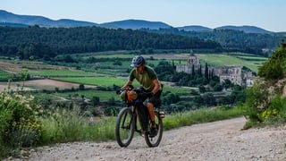 Rider with an orange handlebar bag climbs a white gravel road, a large church and vineyards in the valley behind.