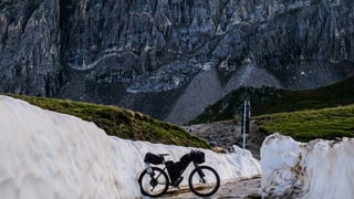 Loaded bikepacking bike parked between snow banks below a sheer rock face on a high mountain road.