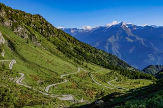 Gravel road zigzagging down a steep green mountainside with snow-capped peaks beyond.