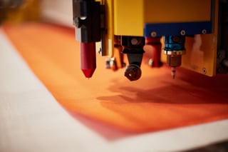 Cutting head of Restrap's automated fabric cutting machine poised over a sheet of orange fabric.