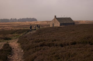 Riders on gravel bikes follow a stony track past an old stone moorland hut on the way down to Ilkley.