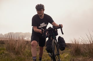 Rider in a 'Bike Packing' t-shirt pushes a gravel bike with bar and fork bags through long moorland grass.