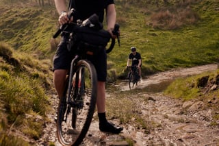 Rider wheels a bike through a muddy ford in the foreground while another splashes across on a moorland byway.