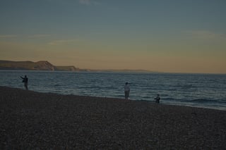 Three figures at the water's edge on the shingle beach at Lyme Regis at dusk, with cliffs along the coast.