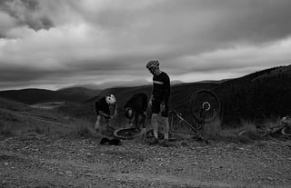 Three riders fix a puncture beside an upturned bike on a hilltop track at Grinduro 2021, in black and white.