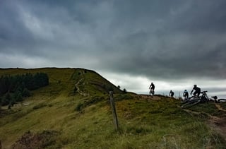 Silhouetted riders crest a grassy ridge under heavy cloud at Grinduro 2021 in Wales, one bike dropped by the path.