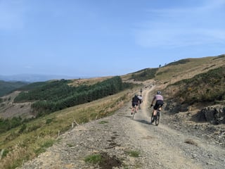 Riders follow a wide stony forestry track up a sunlit Welsh hillside on the Grinduro 2021 course.