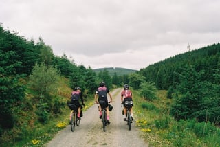 Three Farr Out Deliveries riders pedal a gravel forest road between conifer plantations on the Rapha Pennine Rally.