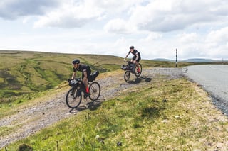 Two Pennine Rally riders ride a stony track beside a moorland road, loaded with bikepacking bags.