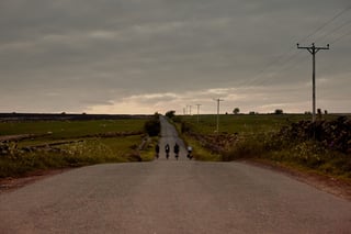 Four bikepackers ride away down a long straight lane between dry stone walls and pastures, heading north from Leeds.