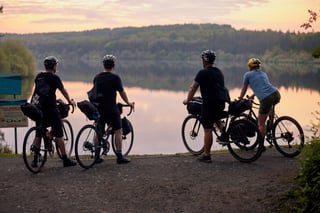 Four riders with loaded gravel bikes pause at a reservoir at dusk during the Restrap factory visit overnighter.