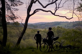 Two riders with loaded bikes look out over a wooded valley at sunset on the Restrap overnighter north of Leeds.
