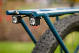 Two small black lights clipped under the blue steel rack of the Clandestine Long Tail, with a knobbly tyre behind.