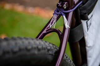 Purple Lord Cycles Nomad frame with a knotted cord on the seatstay bridge beside a chunky tyre and white frame bag.