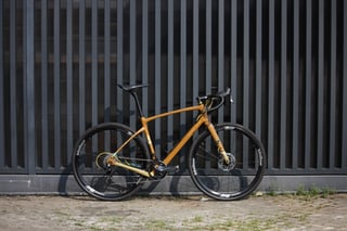 Built-up bronze Chapter2 AO gravel bike with Vision wheels and drop bars standing against a dark slatted fence.