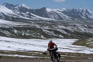 Silk Road Mountain Race rider pushes a loaded bike across snow below mountain peaks in Kyrgyzstan.