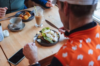 A rider in an orange jersey tucks into an open sandwich with salad and egg at a café table, latte alongside.